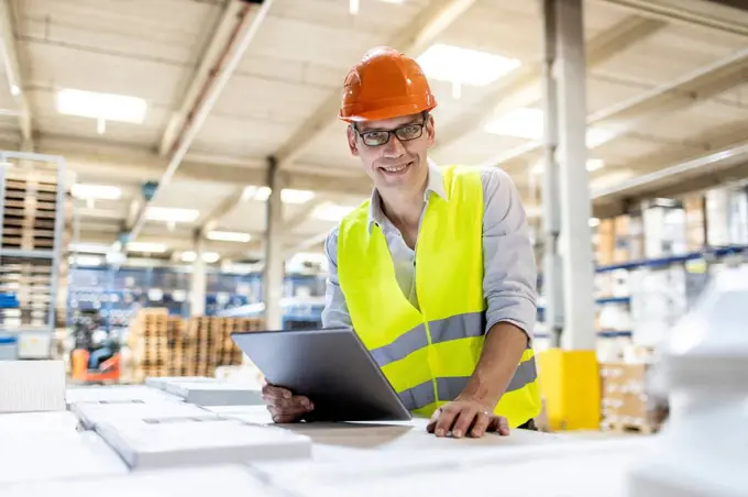 Smiling worker with tablet PC leaning on box in warehouse