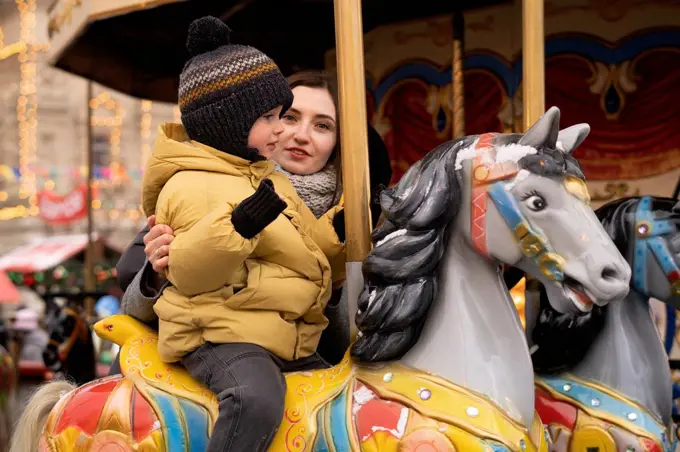 Woman supporting boy sitting on carousel horse in Christmas market