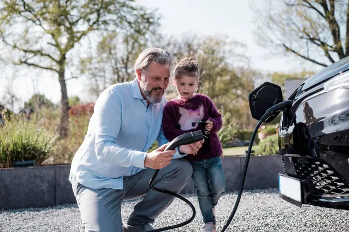 Man showing electric plug to daughter standing in front of car on sunny day