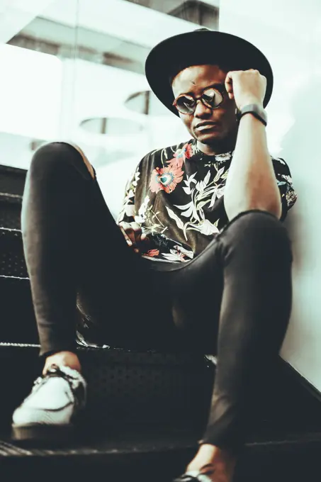Portrait of fashionable man wearing hat, sunglasses and black t-shirt with floral design siitng on stairs
