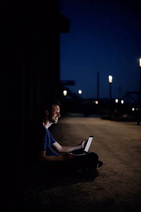 Mid adult man sitting cross-legged on ground, using laptop at night
