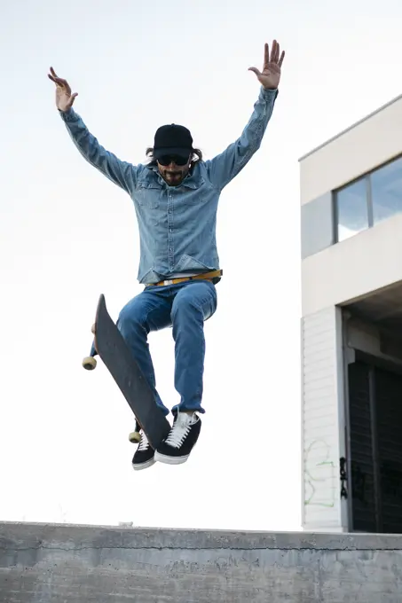 Trendy man in denim and cap skateboarding, doing jump with skateboard from concrete ramp