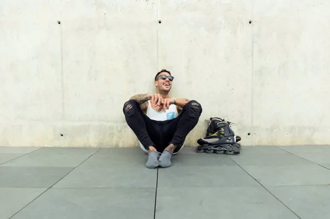 Tattooed young man with roller skates sitting on ground having fun