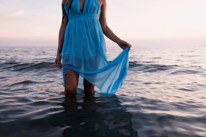 Young tattooed woman wearing blue dress standing in the sea by sunset, partial view