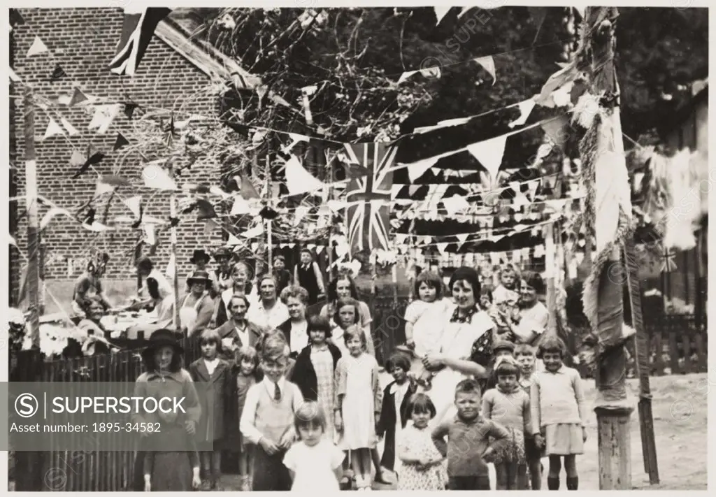A photograph of a street and its residents in Botley, Oxfordshire, taken by an unknown photographer in May 1937. The street has been decorated to cele...