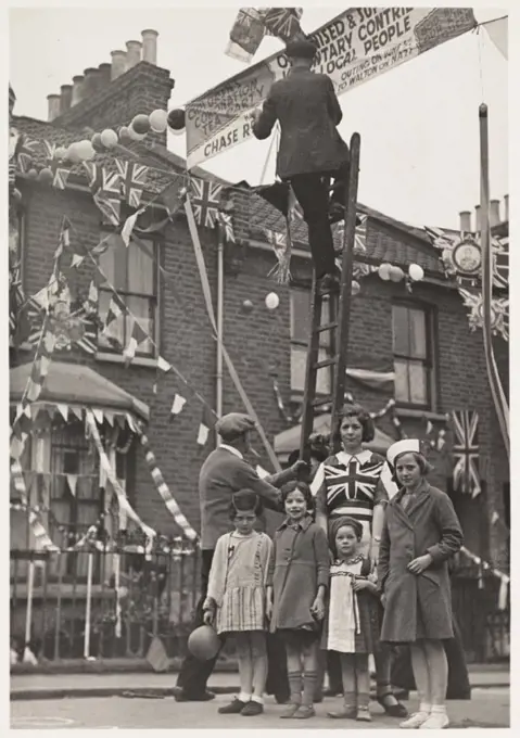 A photograph of residents of a street in North London putting up decorations, taken by an unknown photographer in May 1937. The decorations are to cel...