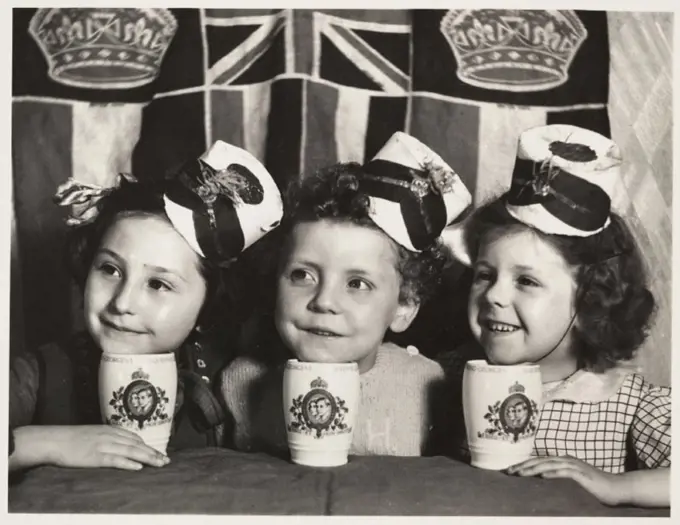 A photograph of three little girls at a Coronation tea party held in Leeds, Yorkshire, taken by an unknown photographer in May 1937. The girls have be...