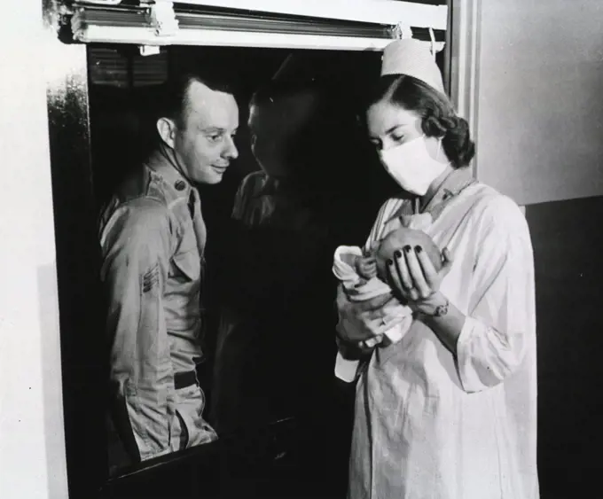 A nurse holds a newborn baby up to the window of a hospital ward. Behind the window, a serviceman in uniform stares at the baby. 