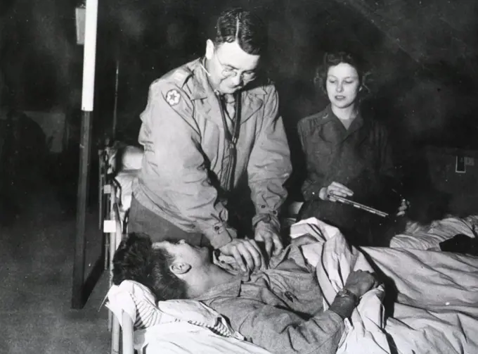 A doctor leans over and unbuttons the shirt of a wounded man lying in bed while a nurse holds a medical chart and watches the patient. 