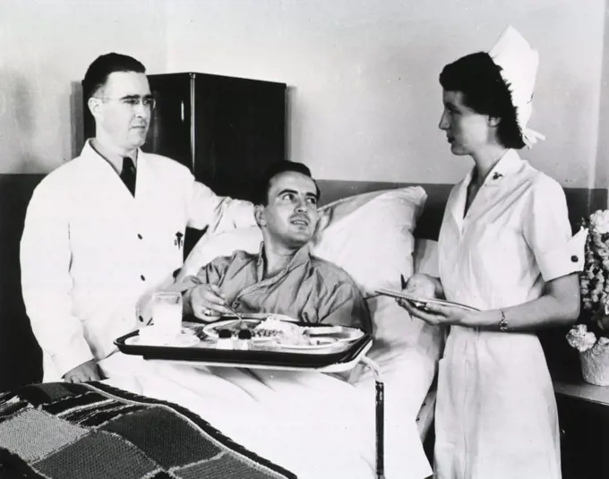 A male doctor and female nurse stand on either side of a hospital bed in which a male patient sits before a food tray. 
