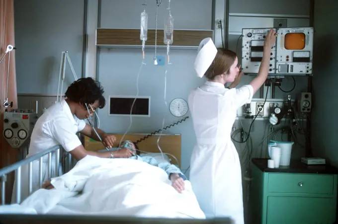 1975 - A nurse examines a patient while another nurse adjusts a monitor. 