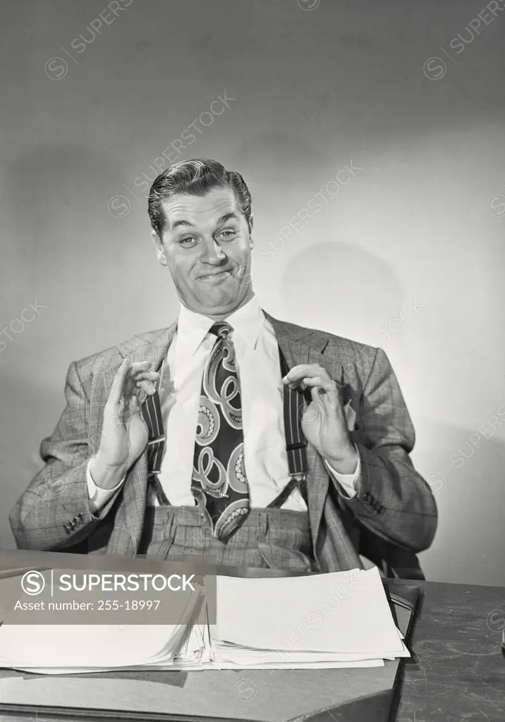 Man in suit and tie sitting behind desk with papers pulling suspenders