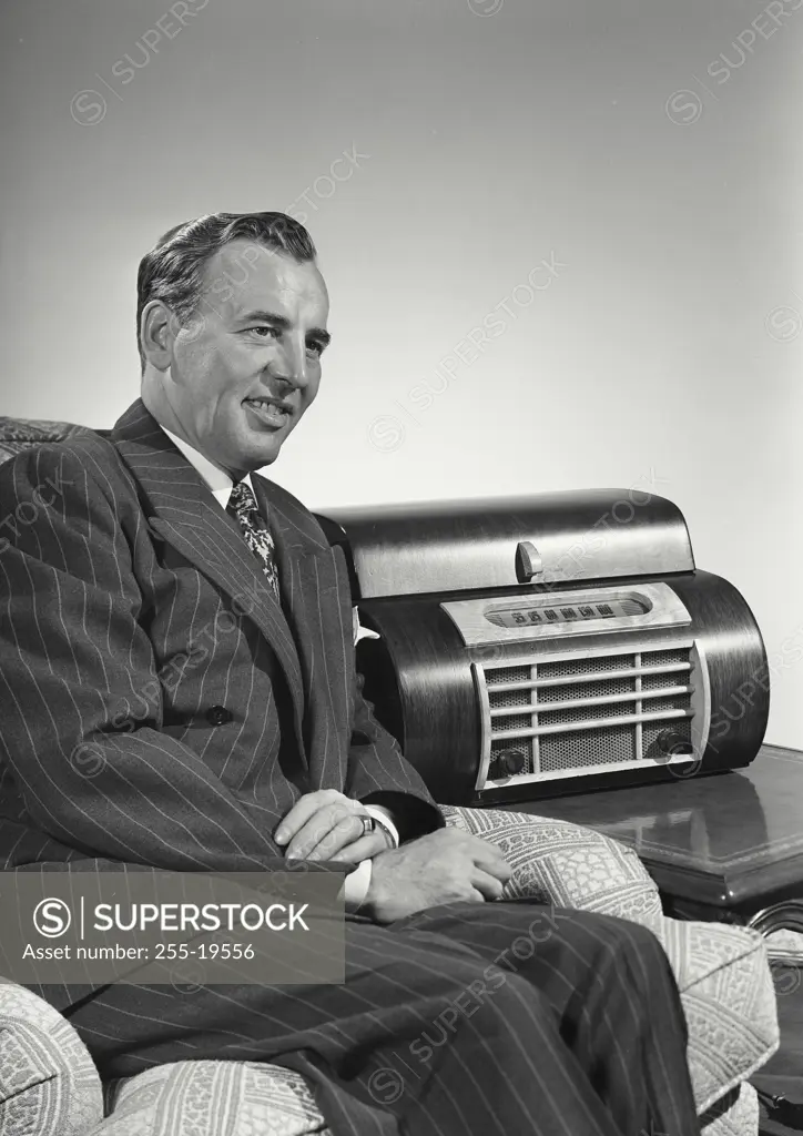 Vintage Photograph. Caucasian man wearing pin striped suit sitting in arm chair next to large radio