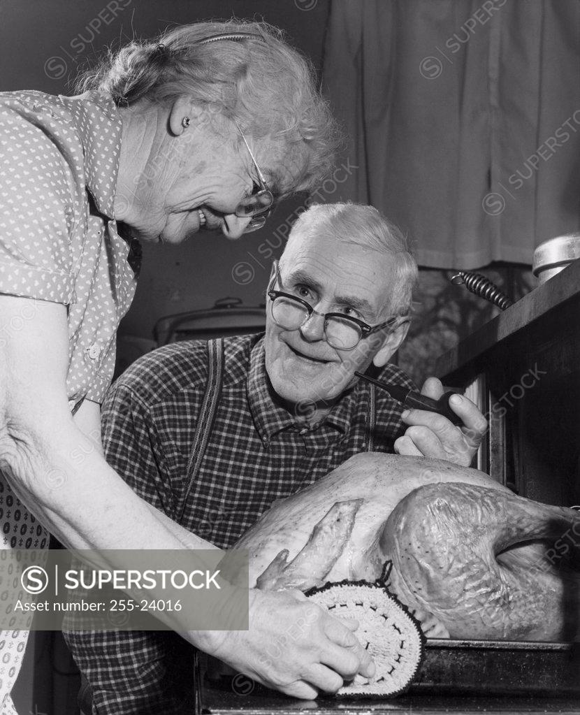 Stock Photo: 255-24016 Side profile of a senior woman putting a turkey into an oven with a senior man looking at her