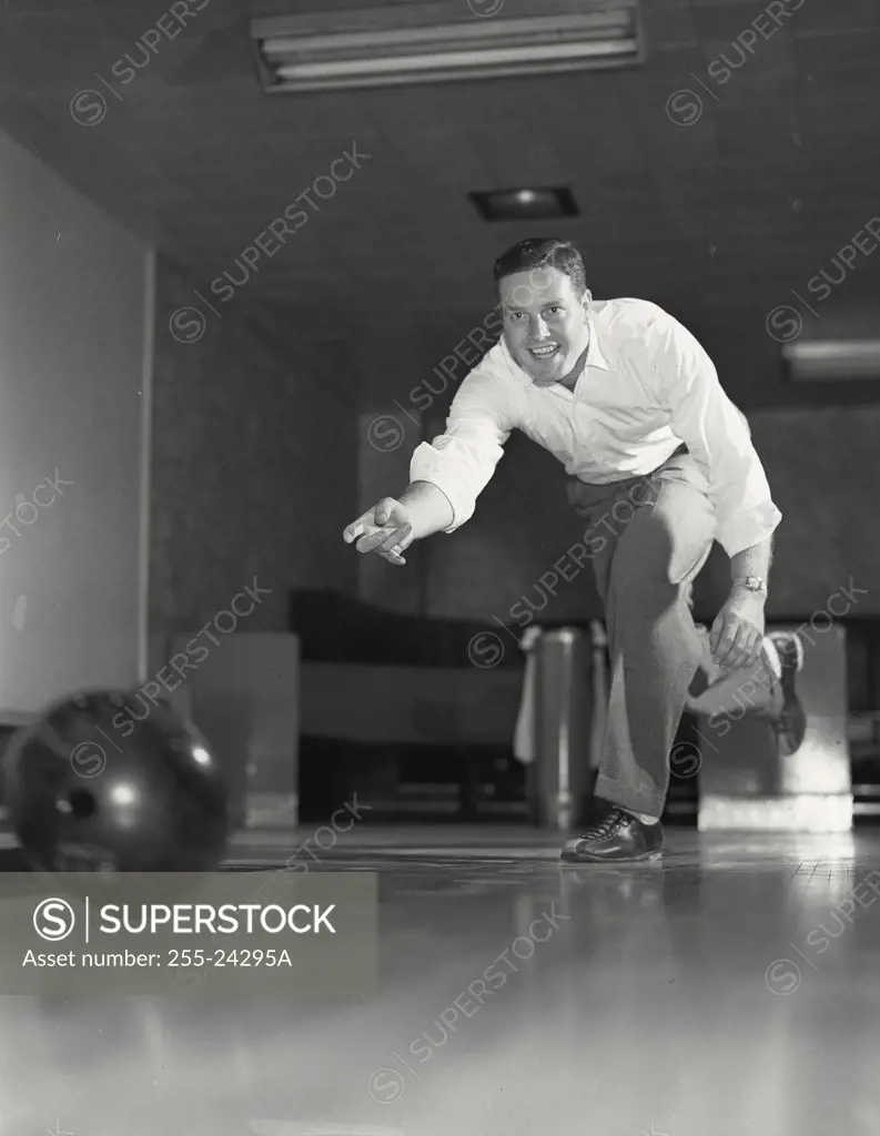 Vintage photograph. Low angle view of a smiling young adult man throwing bowling ball down lane.