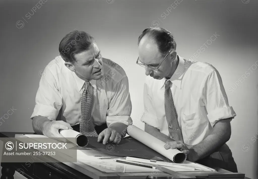 Two men at work desk discussing plans and looking over blueprints.