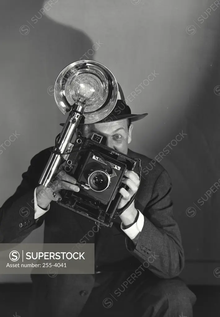 Portrait of press photographer holding up Speed Graphic camera looking through viewfinder at viewer with one eye open