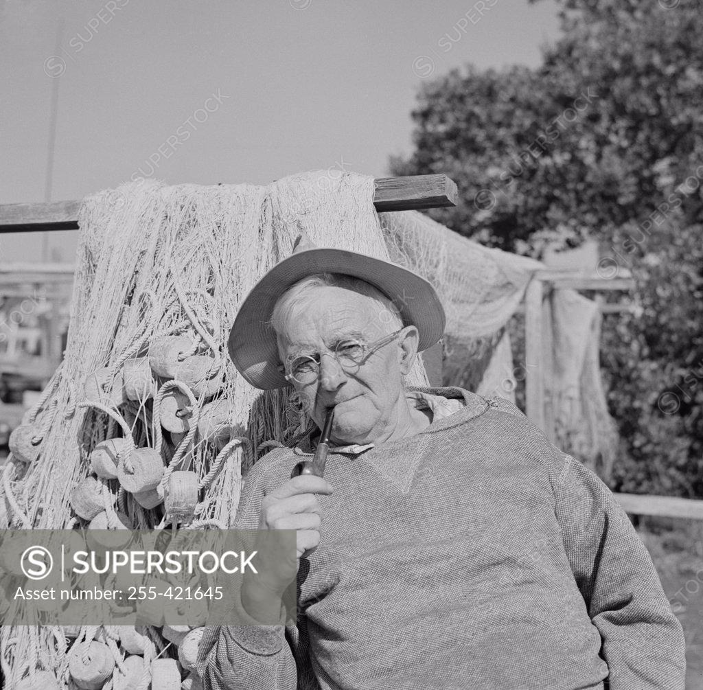 Stock Photo: 255-421645 Senior man wearing hat and smoking pipe, fishing nets in the background