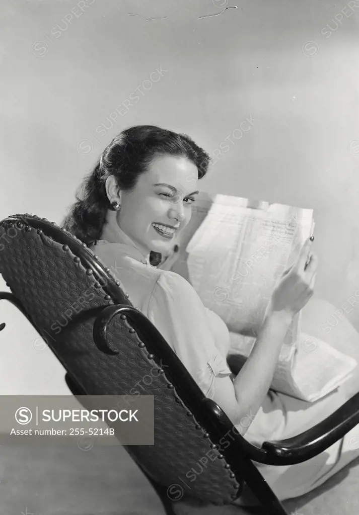 Woman with dark hair sitting in rocking chair holding newspaper winking