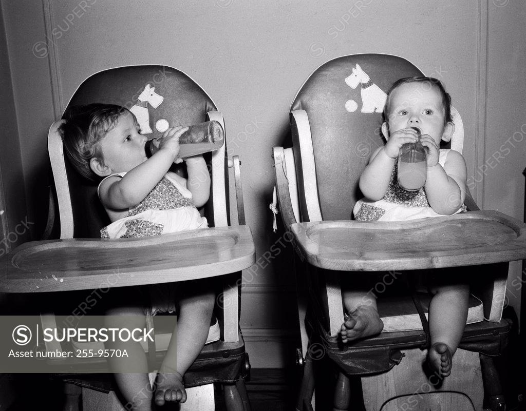 Stock Photo: 255-9570A Two babies sitting in high chairs drinking from baby bottles