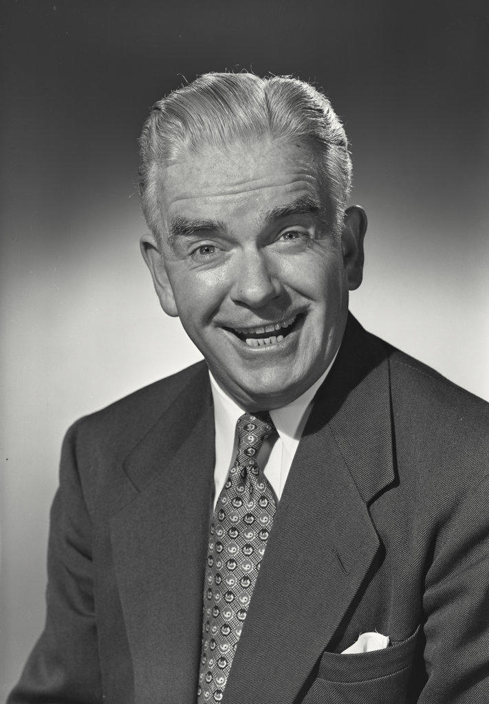 Older man in suit and tie smiling wide at camera with eyebrows raised.