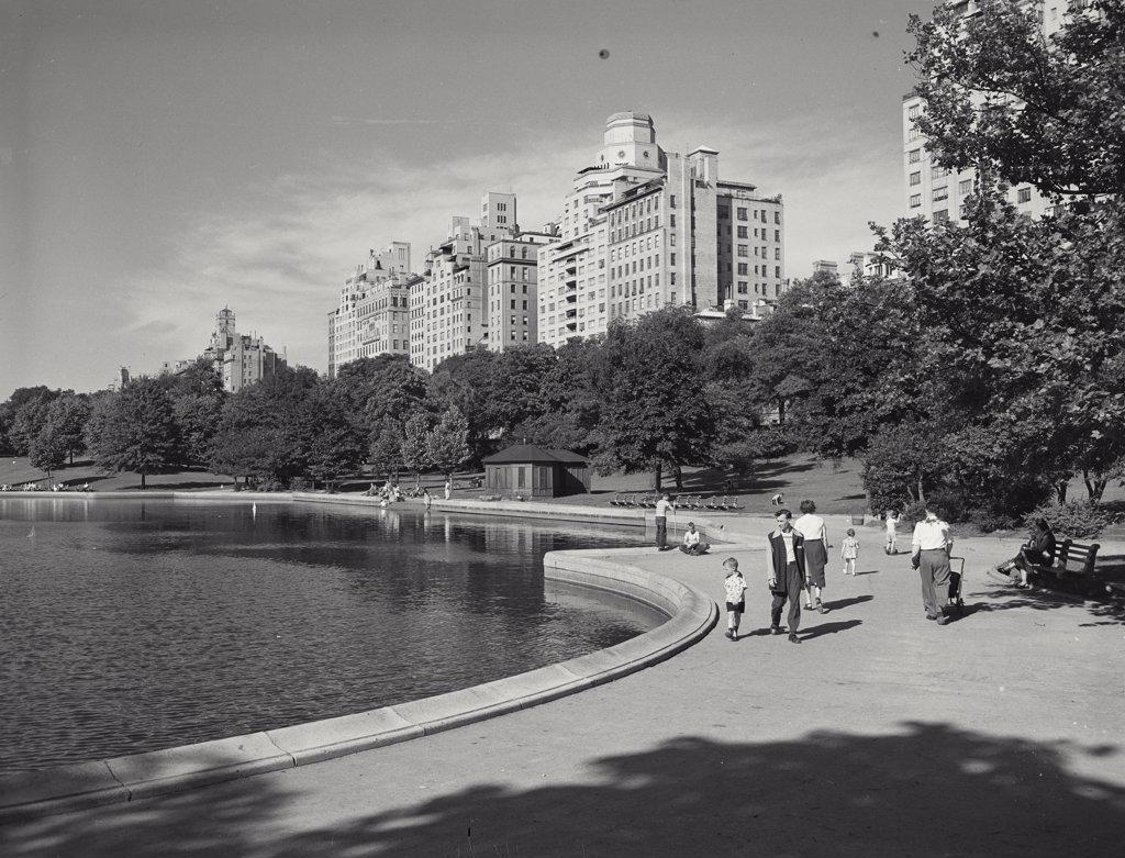 Man and small boy on walkway around pond in Central Park, buildings in background, New York City