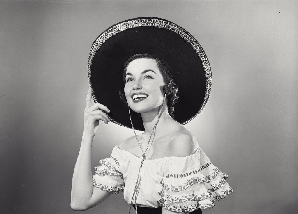 Brunette woman wearing traditional Mexican dress and hand raised to black sombrero with silver trim detail