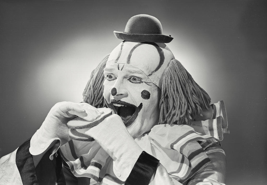 Portrait of clown wearing silly hat with hand sandwiched between two pieces of bread.
