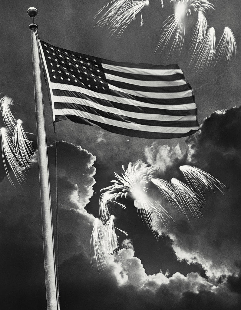 American Flag waving on pole in front of clouds with fireworks