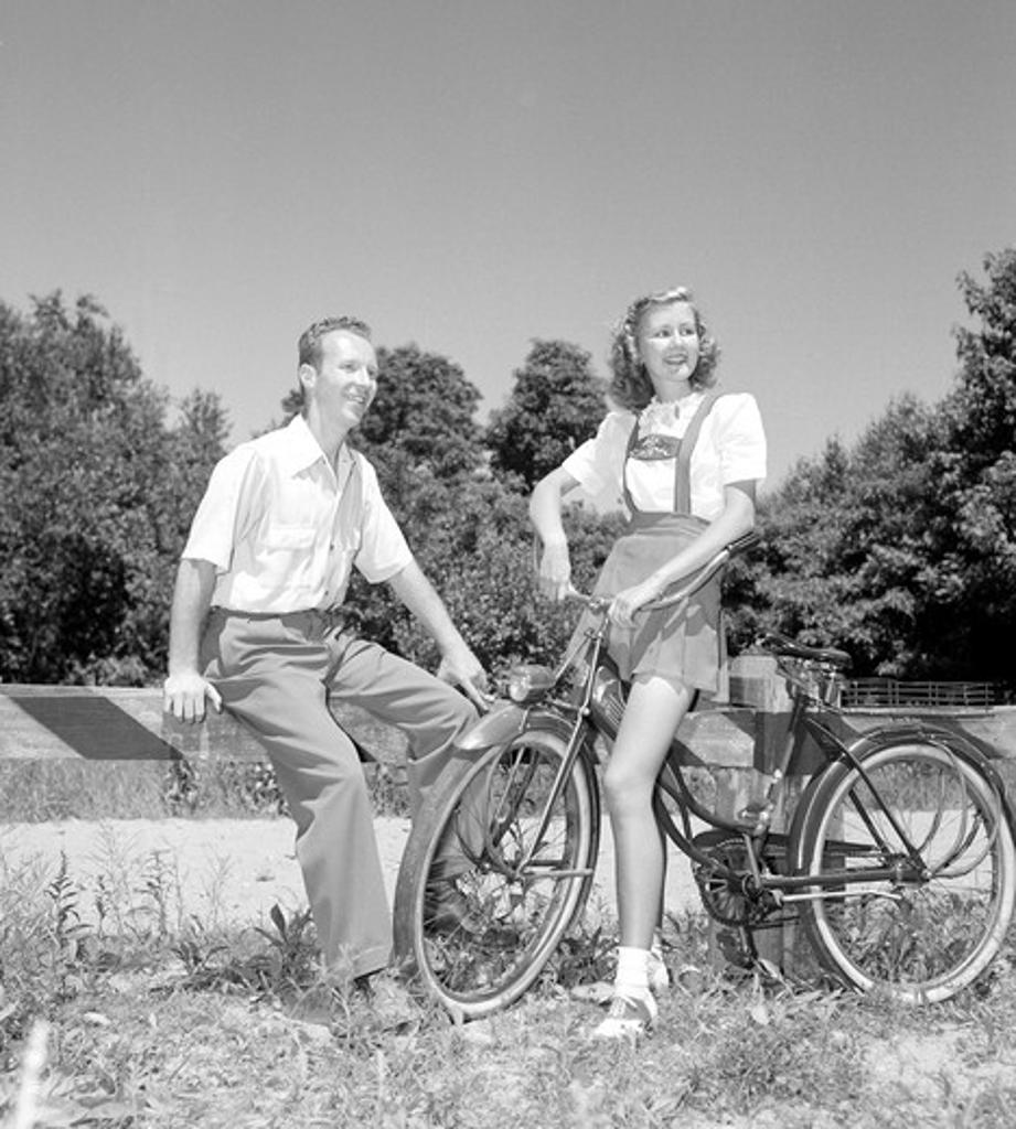 Young man sitting on barrier with young woman standing with bicycle