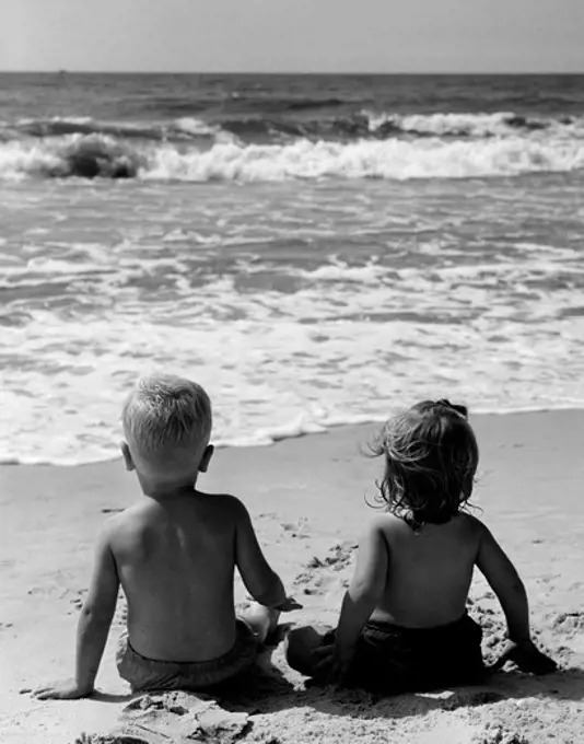 Rear view of pair of children sitting on beach, watching surf