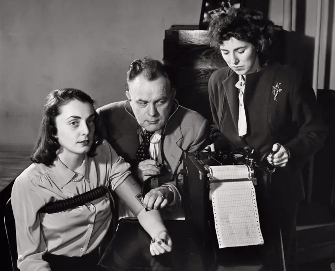 Vintage Photograph. Woman taking polygraph lie detector test while man wearing stethoscope checks her pulse through her am and another woman works the machine