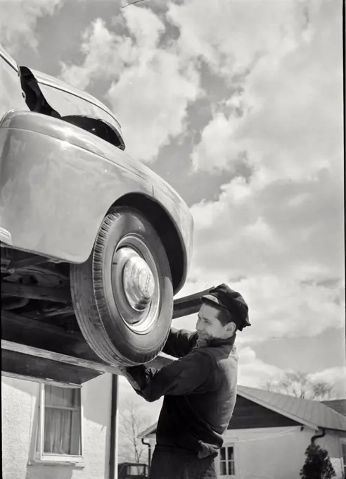 Vintage photograph. Auto mechanic working on car raised up on auto lift outside in Massapequa, Long Island, New York