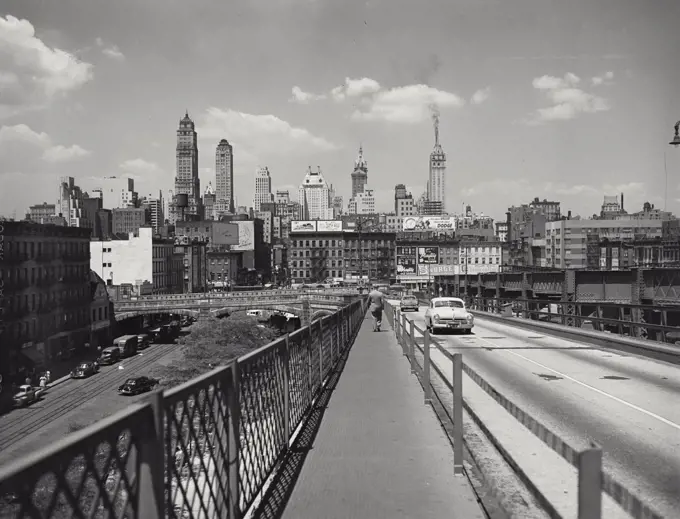Vintage photograph. Man walking on sidewalk of bridge with traffic driving by and skyline in background