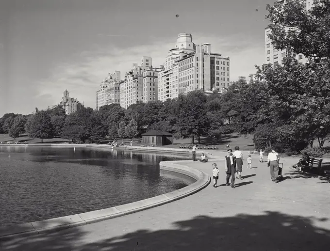 Vintage photograph. Man and small boy on walkway around pond in Central Park, buildings in background, New York City
