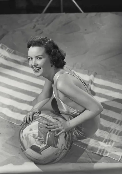 Vintage photograph. Brunette woman smiling wearing bathing suit sitting on beach towel with ball