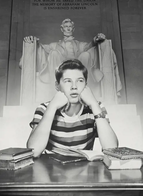 Boy at desk with book open resting cheek on fist with the Lincoln Memorial behind 