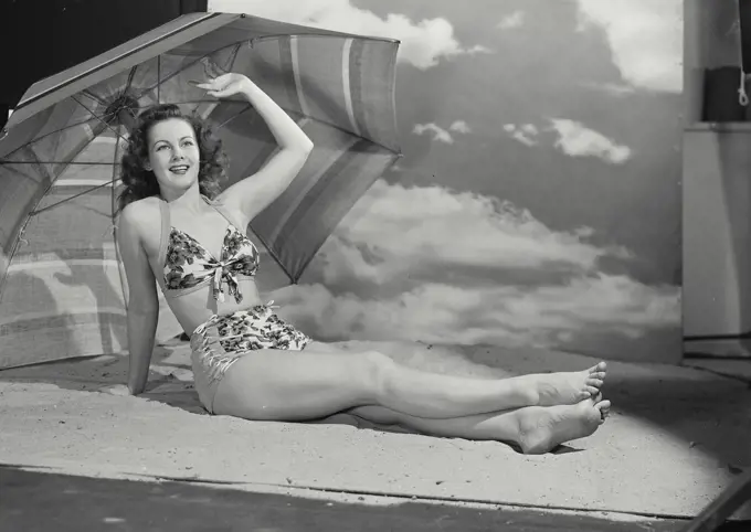 Vintage photograph. Smiling curly haired woman wearing swimsuit lounging with legs outstretched in sand on beach set with large umbrella and sky backdrop with arm raised up