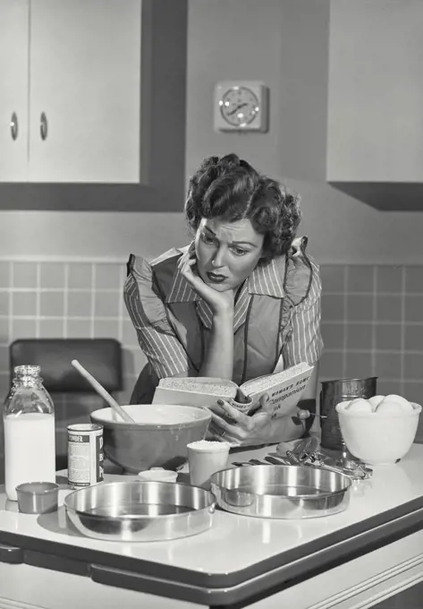 Vintage photograph. Mid adult woman reading a cookbook in a domestic kitchen