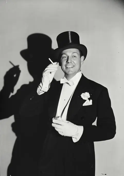 Vintage Photograph. Man in top hat and tuxedo holding cigarette up with shadow behind him