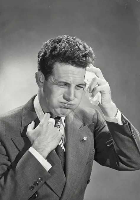 Man with curly dark hair wearing suit and tie holding handkerchief to forehead puffing out cheeks looking uncomfortable