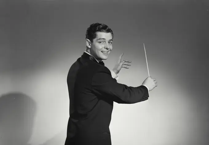 Vintage Photograph. Young man wearing black tuxedo with back turned to viewer holding conductor wand standing in front of solid studio backdrop with light gradient