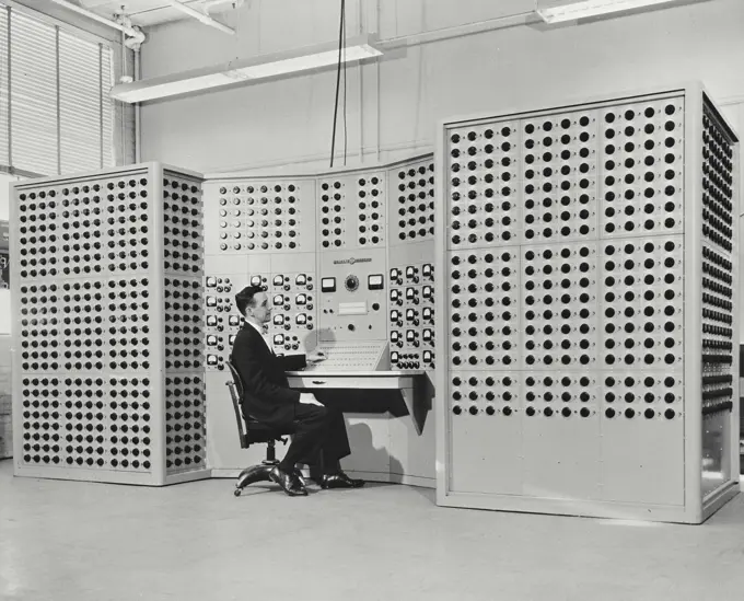 Vintage photograph. Man sitting in front of dial studded new computer that mathematically models a complex electric power system.