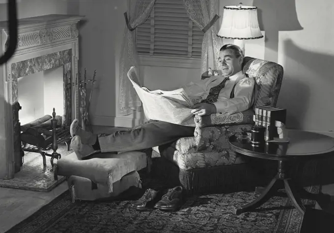 Vintage photograph. Man in chair with feet up next to fireplace reading newspaper