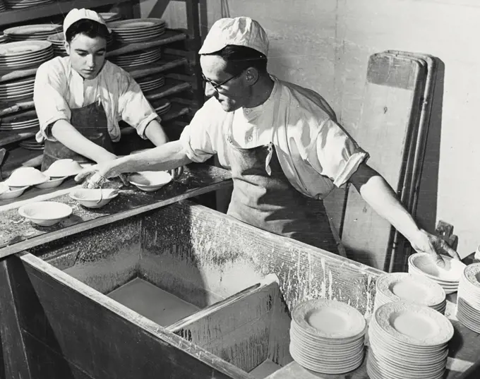 Vintage photograph. British Pottery Industry workers glazing domestic china. The plates are dipped in glaze and then stacked for drying. They wear white caps to protect their heads