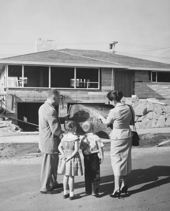 Rear view of a mature couple and their children standing in front of a house and looking at a blueprint