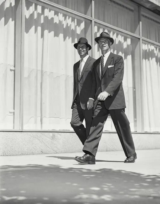 Vintage Photograph. Two men wearing suits and hats walking outside on sidewalk by building with large windows Frame 1