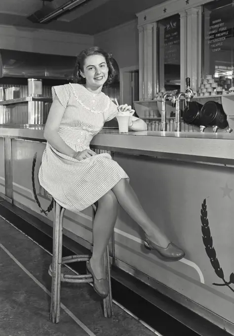 Vintage photograph. Portrait of a teenage girl with milkshake sitting at an ice cream counter and smiling, 1947