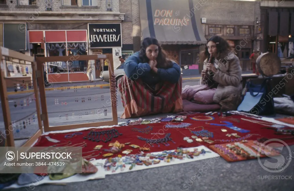 Young woman blowing a flute with another young woman sitting in a market stall, Berkeley, California, USA