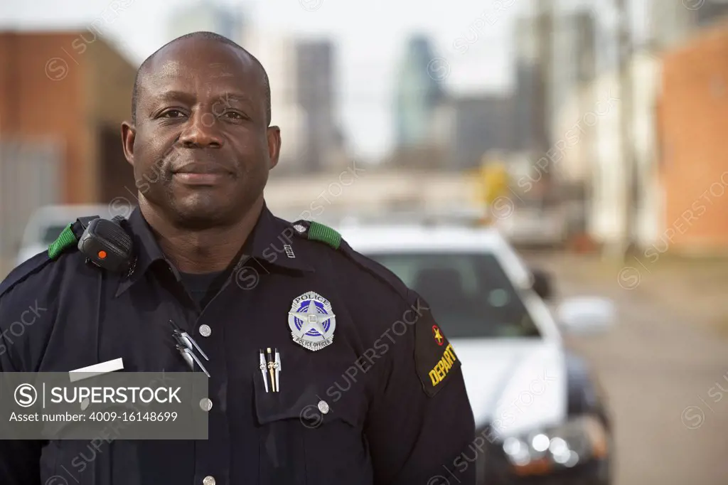 Portrait of Police officer Standing in street in front of squad car  looking towards camera with arms behind his back 